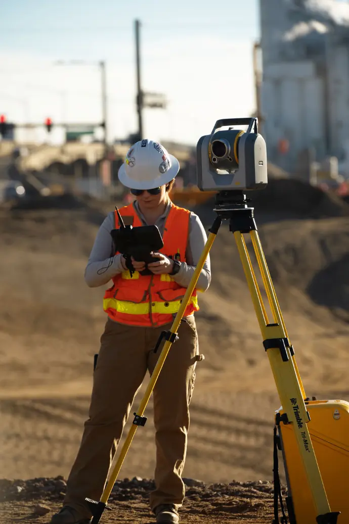 Een landmeter bedient een Trimble SX12 scanning total station op een driepootstatief tijdens meetwerkzaamheden op een bouwplaats.
